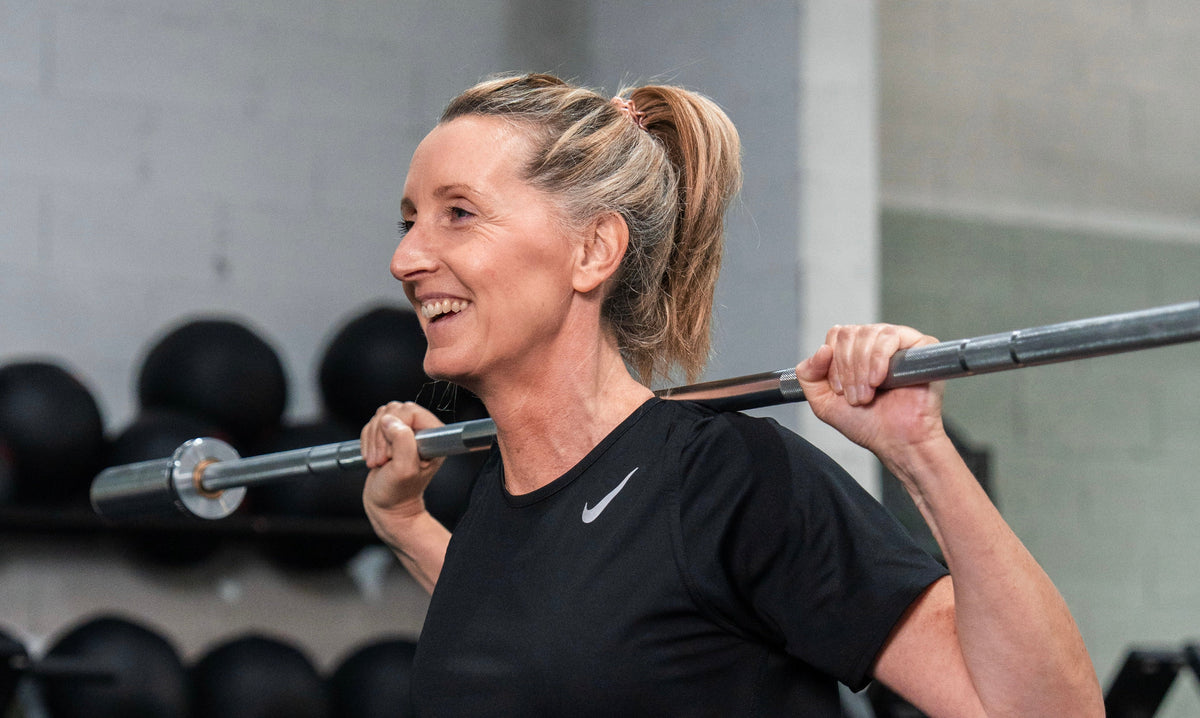 Femme d'une cinquantaine d'années souriante en salle de sport, effectuant un squat avec une barre, illustrant la force fonctionnelle et la préservation de la masse musculaire après 40 ans.